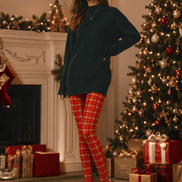 A woman stands in a decorated holiday living room wearing an oversized dark green sweater, red and green plaid tights, and black Mary Jane heels. A Christmas tree, fireplace, and wrapped gifts are visible in the background.