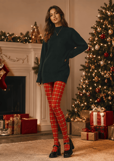 A woman stands in a decorated holiday living room wearing an oversized dark green sweater, red and green plaid tights, and black Mary Jane heels. A Christmas tree, fireplace, and wrapped gifts are visible in the background.