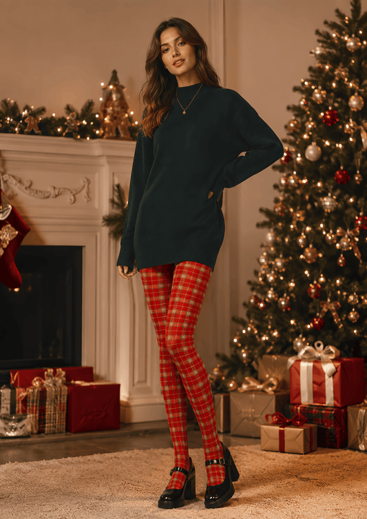 A woman stands in a decorated holiday living room wearing an oversized dark green sweater, red and green plaid tights, and black Mary Jane heels. A Christmas tree, fireplace, and wrapped gifts are visible in the background.